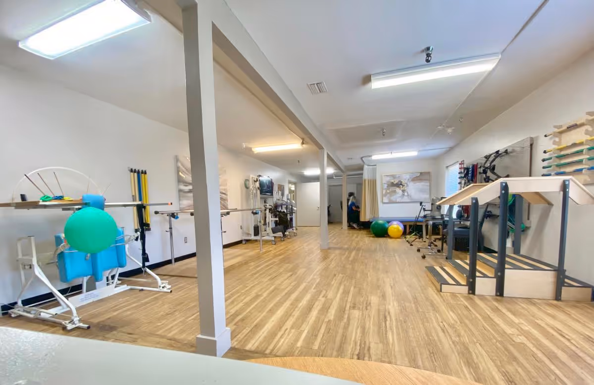 A spacious room with wooden flooring and white walls, equipped with various physical therapy and rehabilitation equipment including parallel bars, exercise balls, and balance training tools. The room is well-lit with ceiling lights and has a calm, clinical atmosphere.