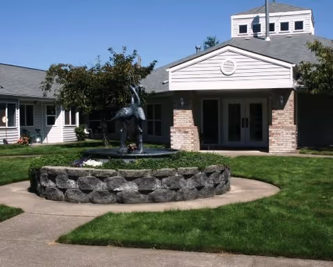 Front entrance of a senior living facility with a circular stone fountain and sculpture in a grassy courtyard.