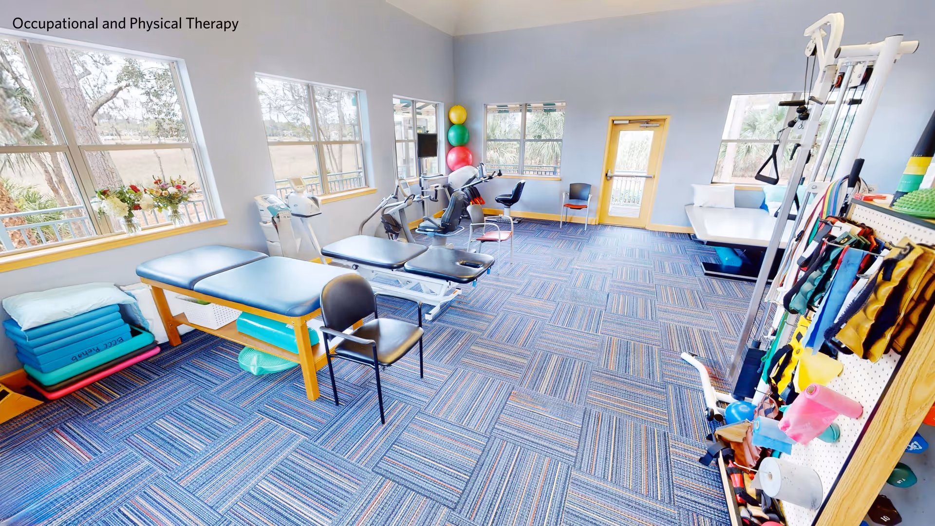 Bright occupational and physical therapy room with treatment tables, exercise equipment, therapy balls and stacked mats near windows.