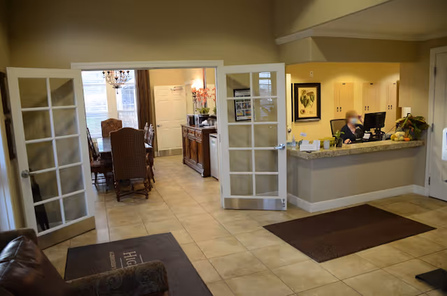 Interior view of a senior living facility reception area with a front desk where a staff member is seated. To the left, there are open double glass doors leading to a dining room with a chandelier and dining table with chairs. The floor is tiled and there are mats on the floor near the entrance and reception desk.