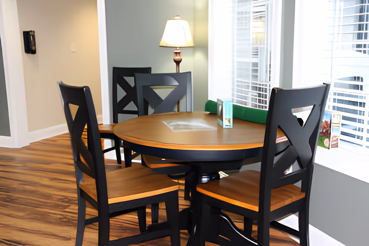 A round wooden dining table with four matching wooden chairs with black frames and wooden seats. The table is placed near a window with white blinds, and a floor lamp with a beige shade is visible in the background. The floor has a wood-like pattern.