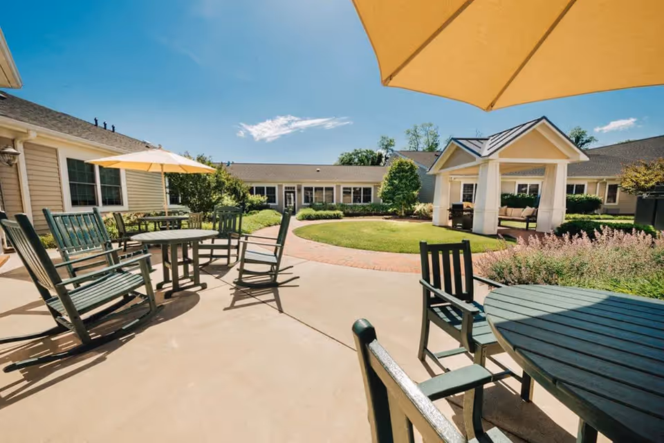 Outdoor patio area at Spring Arbor of Winchester with green wooden chairs and tables, some shaded by yellow umbrellas. There is a circular grassy area with a small pavilion and surrounding buildings under a clear blue sky.