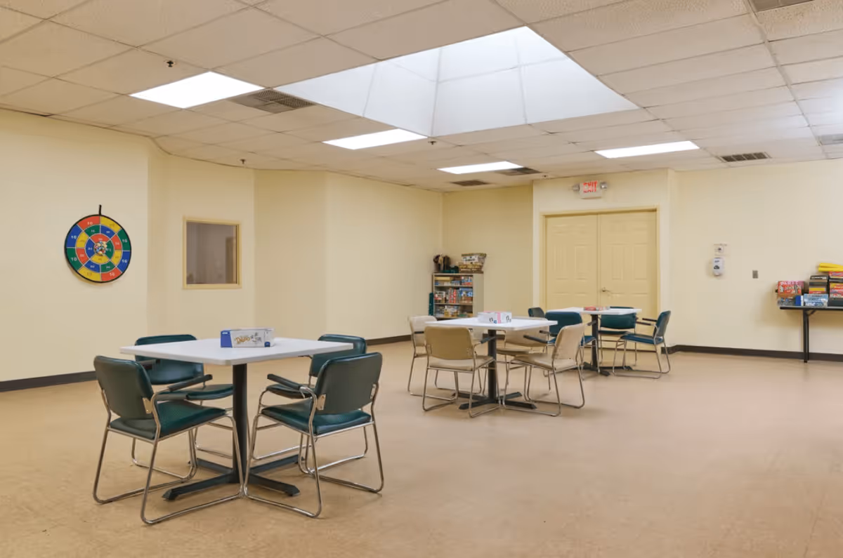 A spacious activity room with several tables and chairs arranged for group activities. The room has a beige floor and cream-colored walls, with a colorful dartboard on one wall and shelves filled with board games in the corner. The ceiling features fluorescent lighting and a skylight.