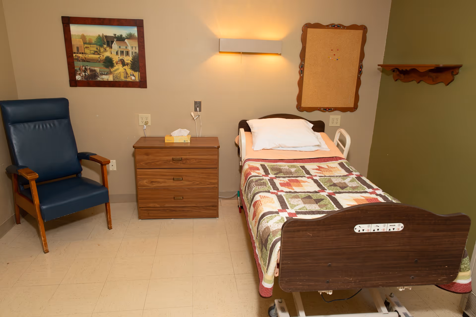 A small room with a single hospital-style bed covered with a patchwork quilt, a white pillow, and a wooden headboard and footboard. Next to the bed is a wooden nightstand with a box of tissues on top. To the left of the nightstand is a blue padded chair with wooden armrests. On the wall above the nightstand is a framed painting depicting a rural scene, and to the right of the bed is a cork bulletin board. The room has beige walls and a tiled floor.
