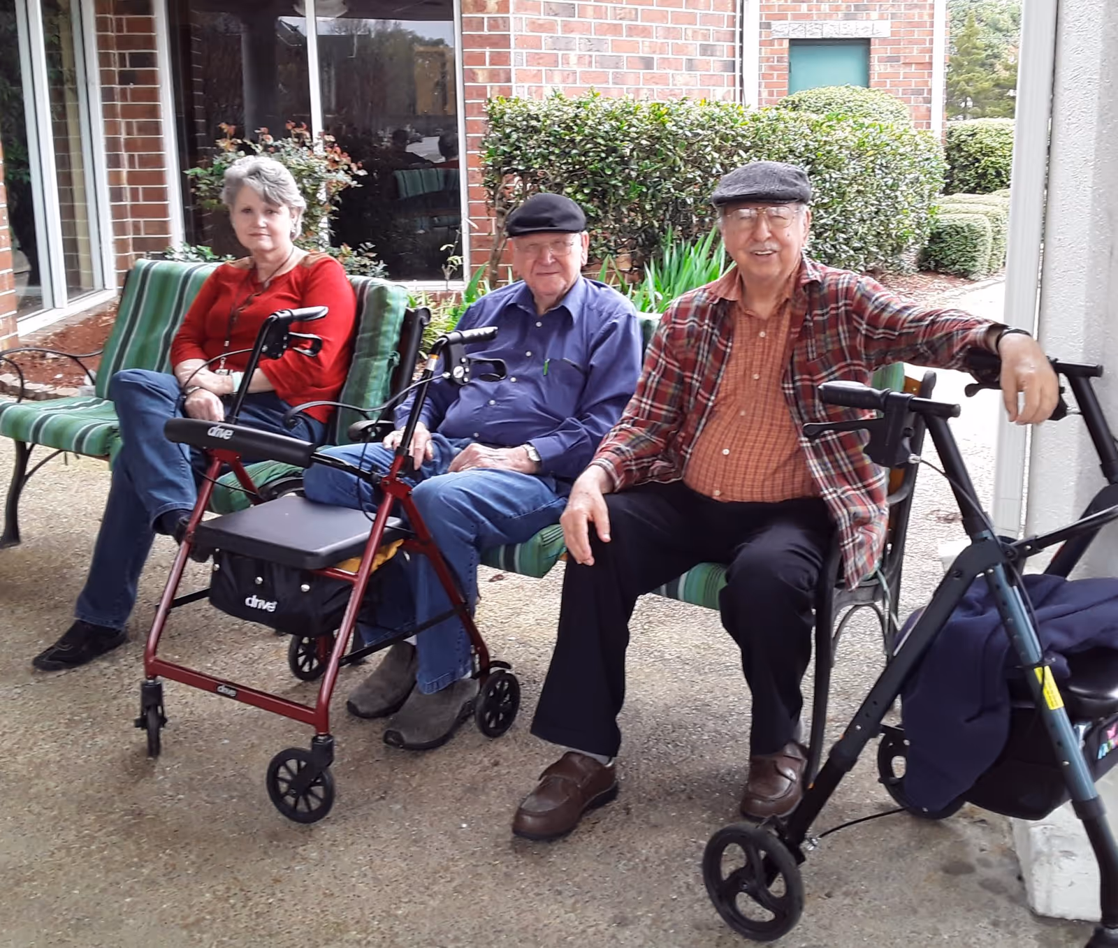 Three elderly individuals sitting on green striped outdoor benches near a brick building. Two men and one woman are seated, each with a walker in front of them. There are bushes and plants in the background.