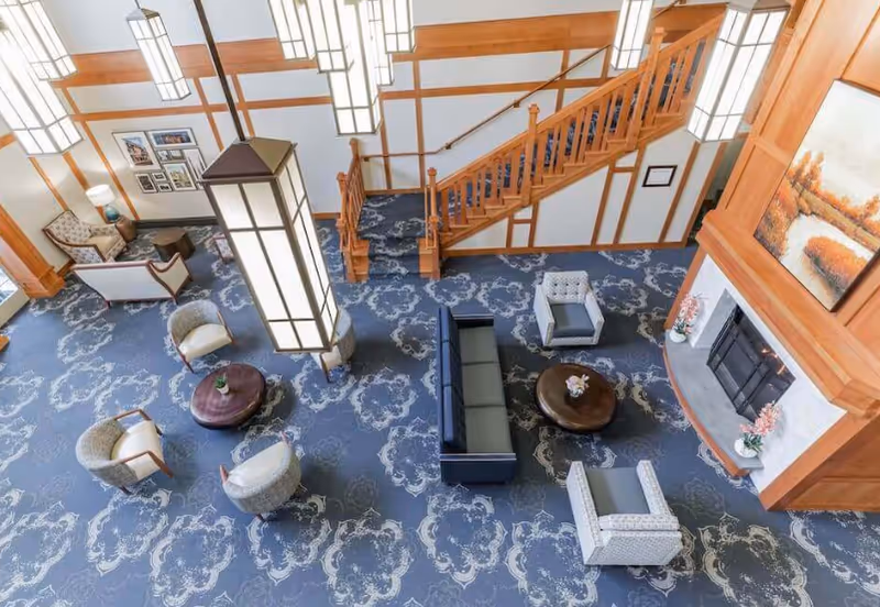 A spacious and well-lit common area with patterned blue carpet, several armchairs and sofas arranged around two round wooden coffee tables, a large wooden staircase, and a fireplace with a painting above it. The room features tall, rectangular hanging light fixtures and wood paneling on the walls.