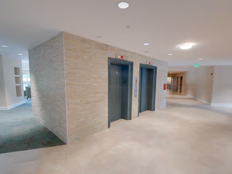 Interior view of a hallway in Linden Park Apartments in Bolton Hill featuring two closed elevators with dark frames set in a beige stone wall. The floor is tiled in light beige, and the hallway extends to the right with neutral-colored walls and ceiling lights. To the left, there is a carpeted area with green patterned carpet and built-in shelving.