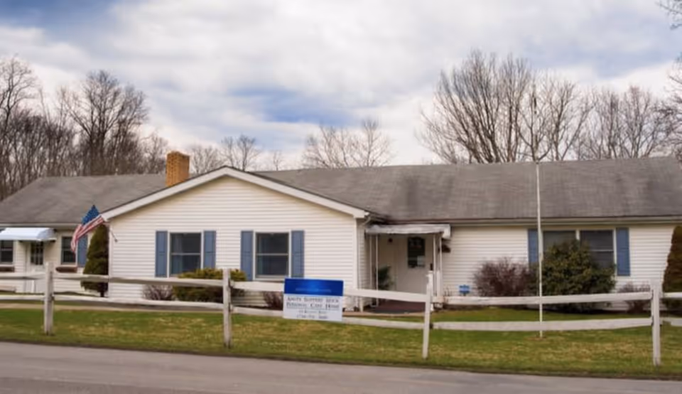 Single-story white building with blue shutters and a gray roof, surrounded by a wooden fence and some bushes. An American flag is displayed near the entrance. The sky is cloudy and leafless trees are visible in the background.