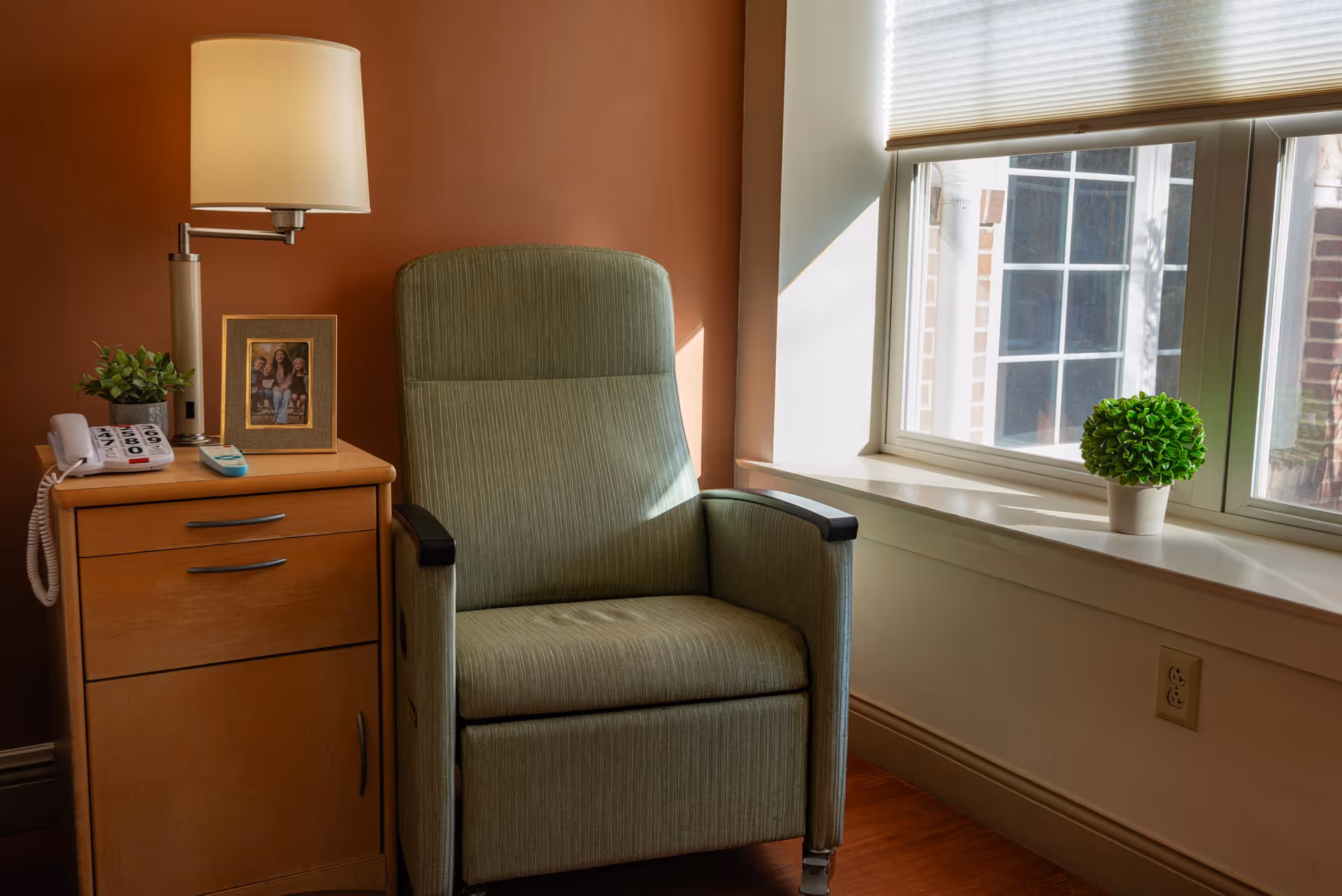 Sunlit corner of a room with a green upholstered recliner beside a wooden nightstand holding a lamp, telephone, framed photo and a small plant by a window.