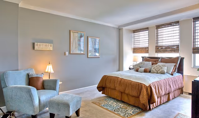 A well-lit bedroom with a neatly made bed, decorative pillows, an upholstered armchair and ottoman, lamps, wall art and three windows with woven shades.