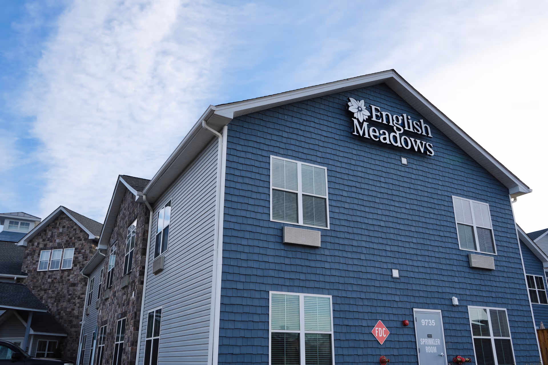 Exterior view of a multi-story building with blue siding and stone accents. The building has several windows and a sign near the roof that reads 'English Meadows'. The sky is partly cloudy.
