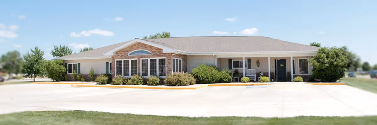 Single-story building with a brick and siding exterior, large windows, and a covered porch with chairs, surrounded by bushes and trees under a clear blue sky.