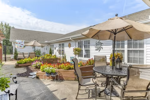 Outdoor patio area at Brookdale Reidsville with a round metal table and four chairs under a large beige umbrella. Raised garden beds with colorful flowers and plants line the side of the building. The building has white siding and multiple windows with white blinds. The sky is clear with some clouds.