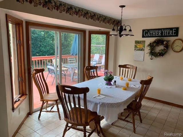 Dining area with a rectangular table covered with a white lace tablecloth, surrounded by six wooden chairs. On the table are six glasses of orange juice and a small flower arrangement. Large sliding glass doors open to a wooden deck with outdoor seating and greenery visible outside. A wall decoration with the words 'Friends & Family Gather Here', a wreath, a clock, and a calendar are on the wall.