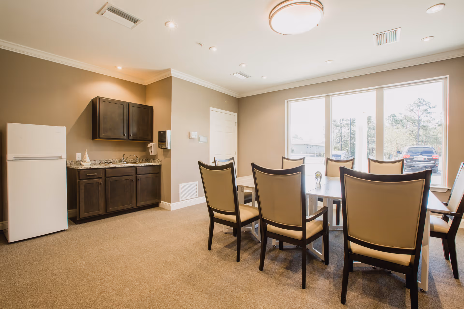 A bright dining area in a senior living facility with a rectangular table surrounded by eight cushioned chairs. The room has large windows letting in natural light, beige walls, and a small kitchenette with dark wood cabinets, a white refrigerator, and a granite countertop.
