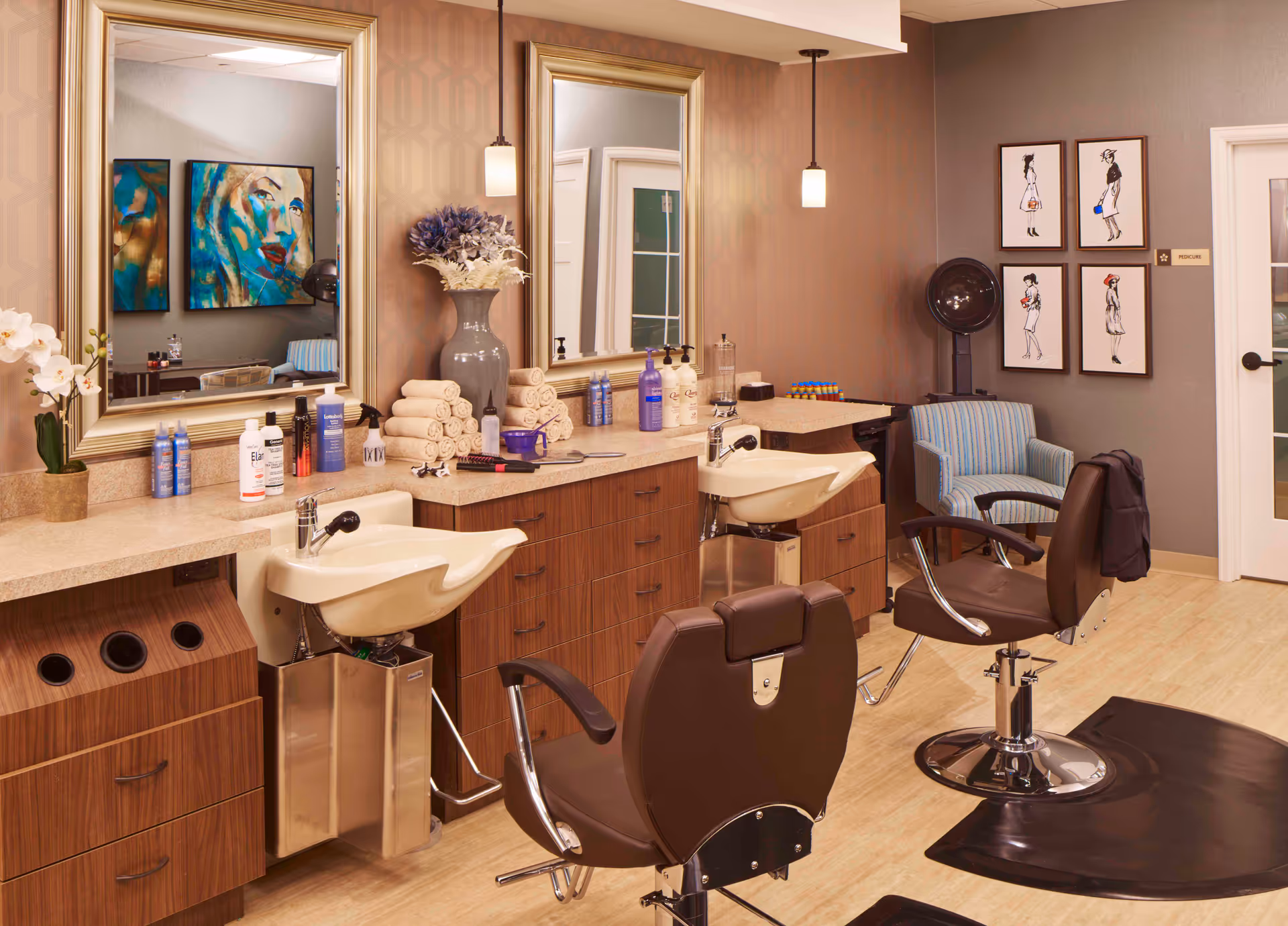 Interior of a senior living facility salon area with two brown salon chairs in front of sinks and mirrors. The countertop holds various hair care products, rolled towels, and a vase with flowers. On the wall are four framed fashion sketches and a sign labeled 'PEDICURE'. A striped armchair and a hair dryer are also visible.