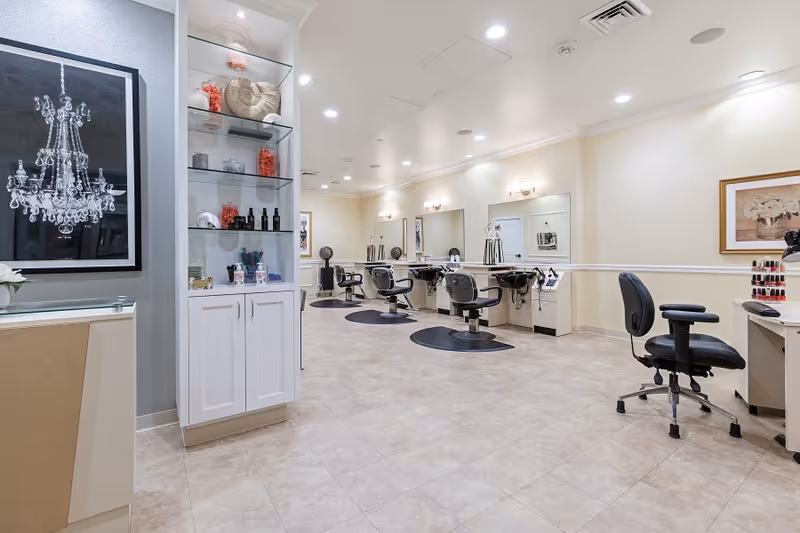 Interior view of a salon area in a senior living facility with multiple black salon chairs in front of mirrors and sinks, a display cabinet with decorative items, and a desk with nail polish bottles. The room has tiled flooring, light-colored walls, and ceiling lights.