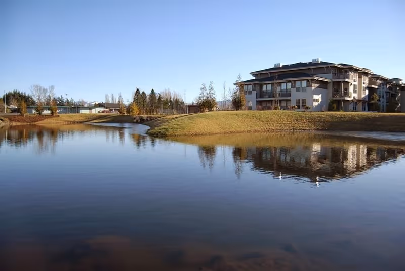 A large pond with clear water reflecting a multi-story residential building and surrounding trees under a clear blue sky.