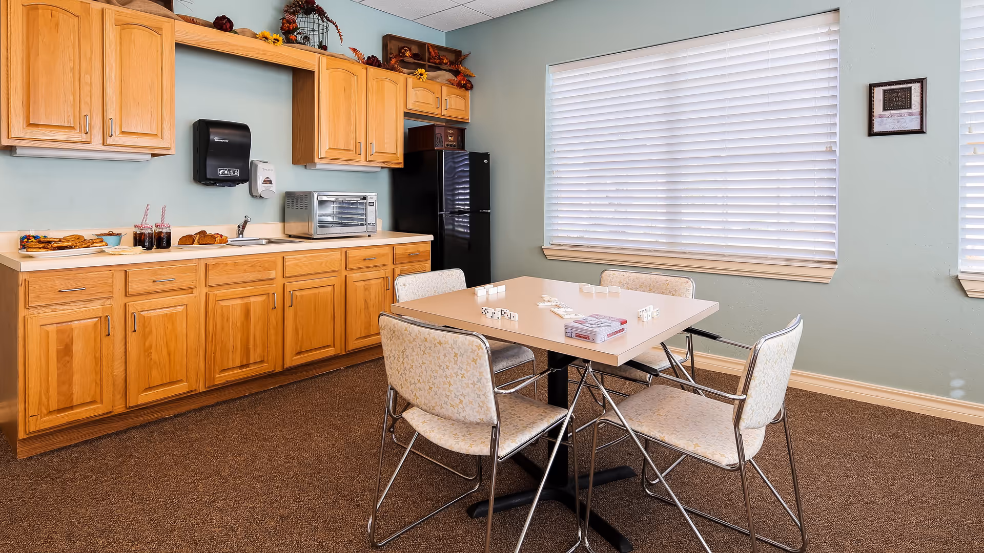 A small kitchen area with wooden cabinets, a countertop with a toaster oven, a sink, and a black refrigerator. A table with four chairs is in the center of the room, with a game of dominoes set up on the table. The room has light blue walls and a large window with closed blinds.