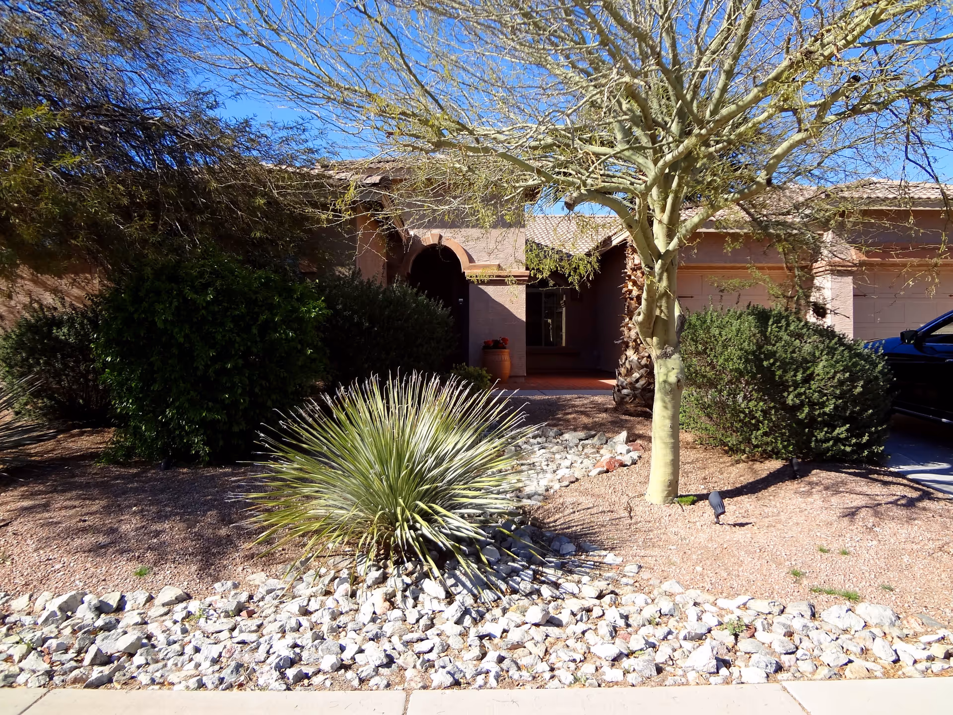 Front exterior view of a single-story residential building with a tiled roof, an arched entrance, and a landscaped front yard featuring desert plants, rocks, and a tree. A black vehicle is partially visible on the right side in the driveway.