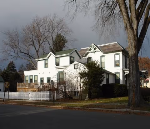 White multi-story senior home on a street with trees, a wooden deck, and a fence.