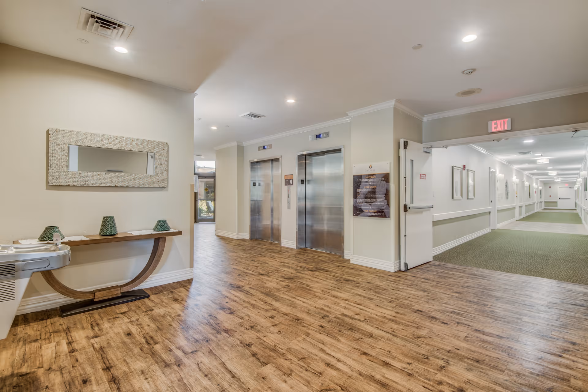 Lobby area with wood flooring, two stainless steel elevators, a console table and a long hallway leading to rooms.