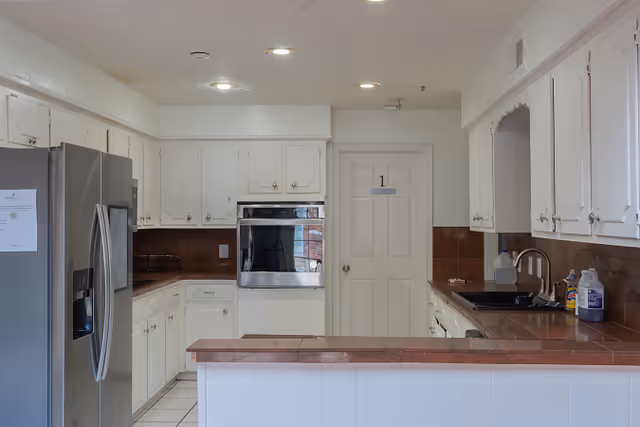 A clean kitchen with white cabinets and brown countertops. There is a stainless steel refrigerator on the left, a built-in oven in the center, and a sink with cleaning supplies on the right. The kitchen has recessed lighting and a white door labeled with the number 1.
