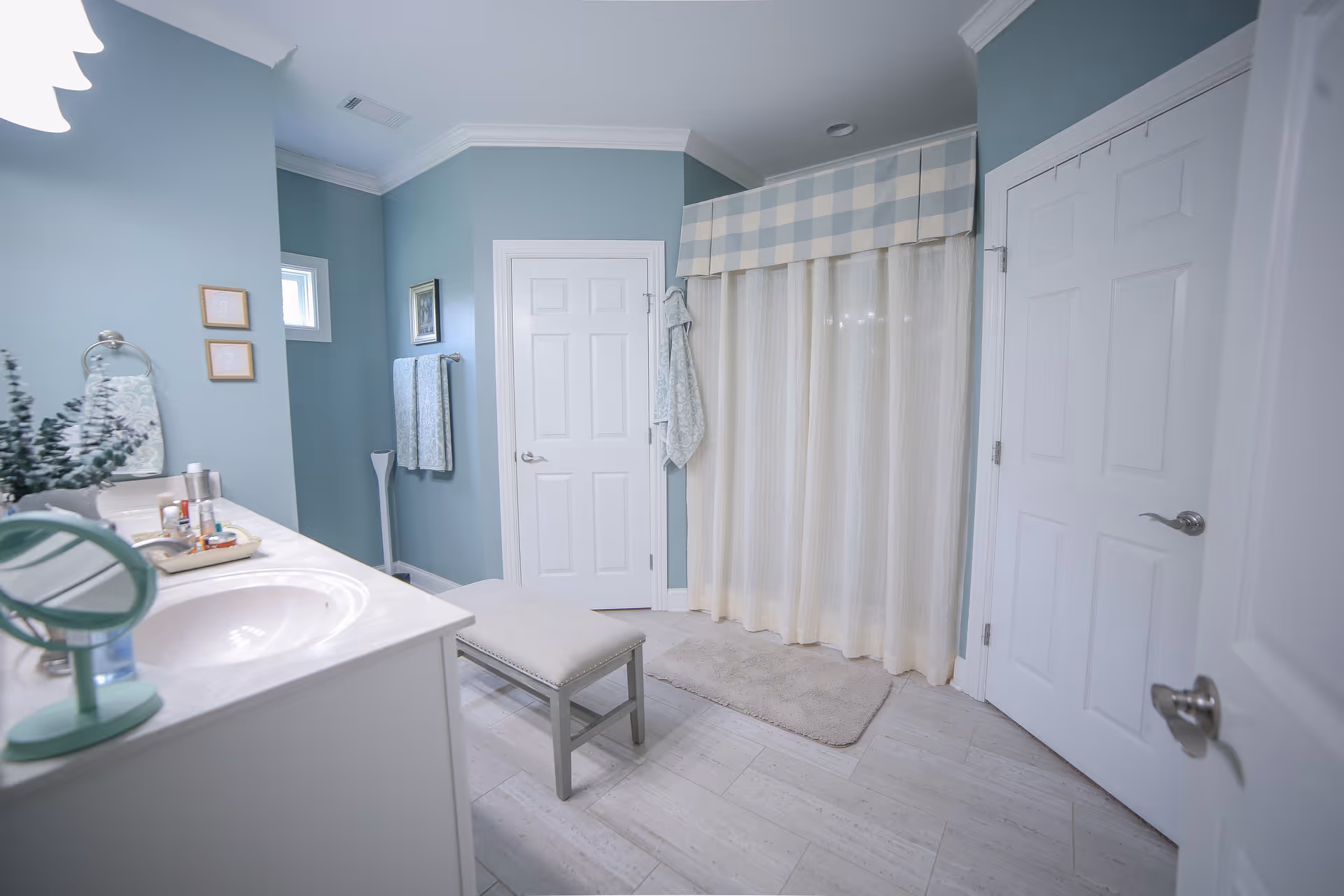 A clean and spacious bathroom with light blue walls and white trim. There is a white vanity with a sink and various toiletries on the left side, a small cushioned bench in the center, and a shower area with a checkered blue and white valance and cream-colored curtains. Two white doors are visible, one on the back wall and one on the right side. Towels hang on a rack near the back wall, and a small window is above the towel rack.