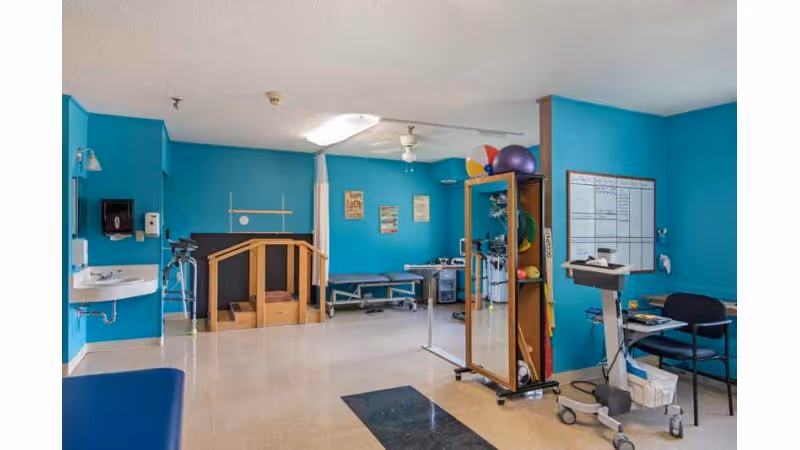 A rehabilitation therapy room with bright turquoise walls, featuring exercise equipment, parallel bars, a small staircase with handrails, therapy balls on a shelf, a sink, a chair, and a whiteboard on the wall.