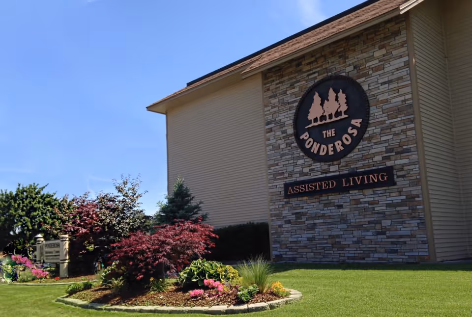 Exterior view of The Ponderosa Assisted Living building with a stone facade and signage. The building is surrounded by a well-maintained lawn and landscaped garden with various shrubs and flowers under a clear blue sky.