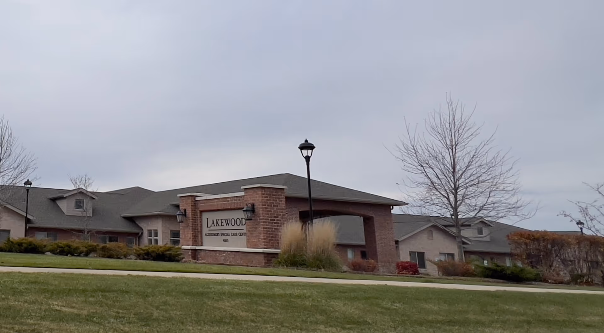 Exterior view of Lakewood Memory Care & Assisted Living facility showing a brick sign with the facility name, surrounded by grass, shrubs, and leafless trees under a cloudy sky.