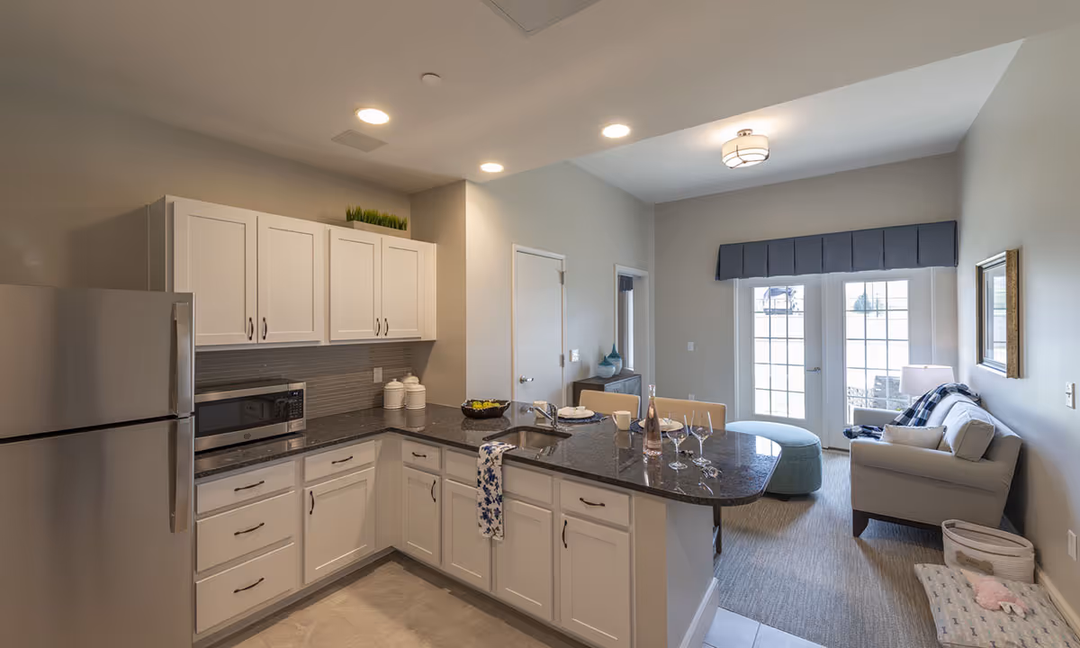A modern open-concept kitchen and living area in an assisted living facility. The kitchen features white cabinets, a stainless steel refrigerator, a microwave, and a dark granite countertop with a sink and a few place settings. The living area has a light gray sofa, a blue ottoman, a side table with a lamp, and large windows with blue valances letting in natural light.
