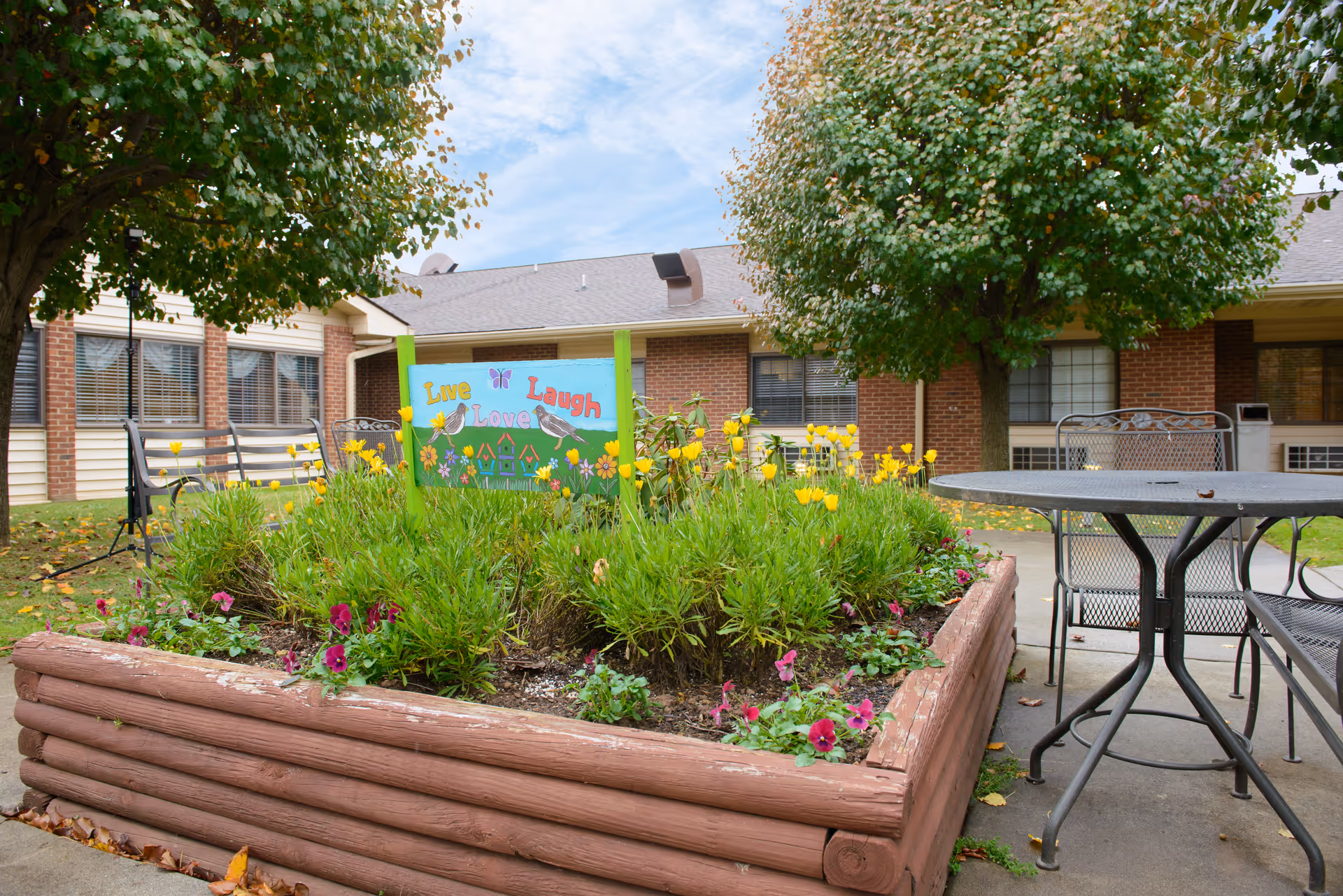 Courtyard garden with a raised flower bed and a colorful "Live Laugh Love" sign, patio table and chairs, and a brick-faced building in the background.