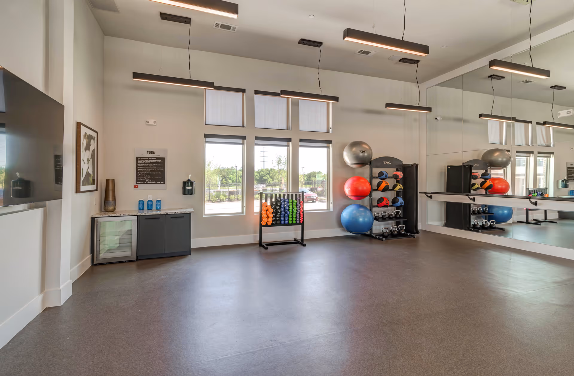 A bright and spacious exercise room with large windows letting in natural light. The room features a rack of colorful dumbbells, exercise balls, kettlebells, and a large wall mirror. Modern hanging lights illuminate the space, and there is a small counter with water bottles and a mini fridge beneath a yoga instruction sign.