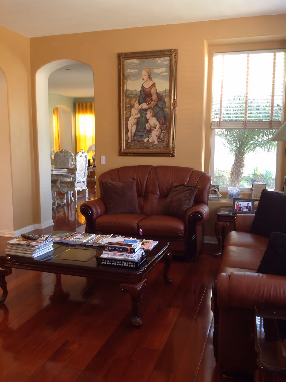 Cozy living room with leather sofas, a wooden coffee table piled with magazines, hardwood floors, and a framed painting above the couch.