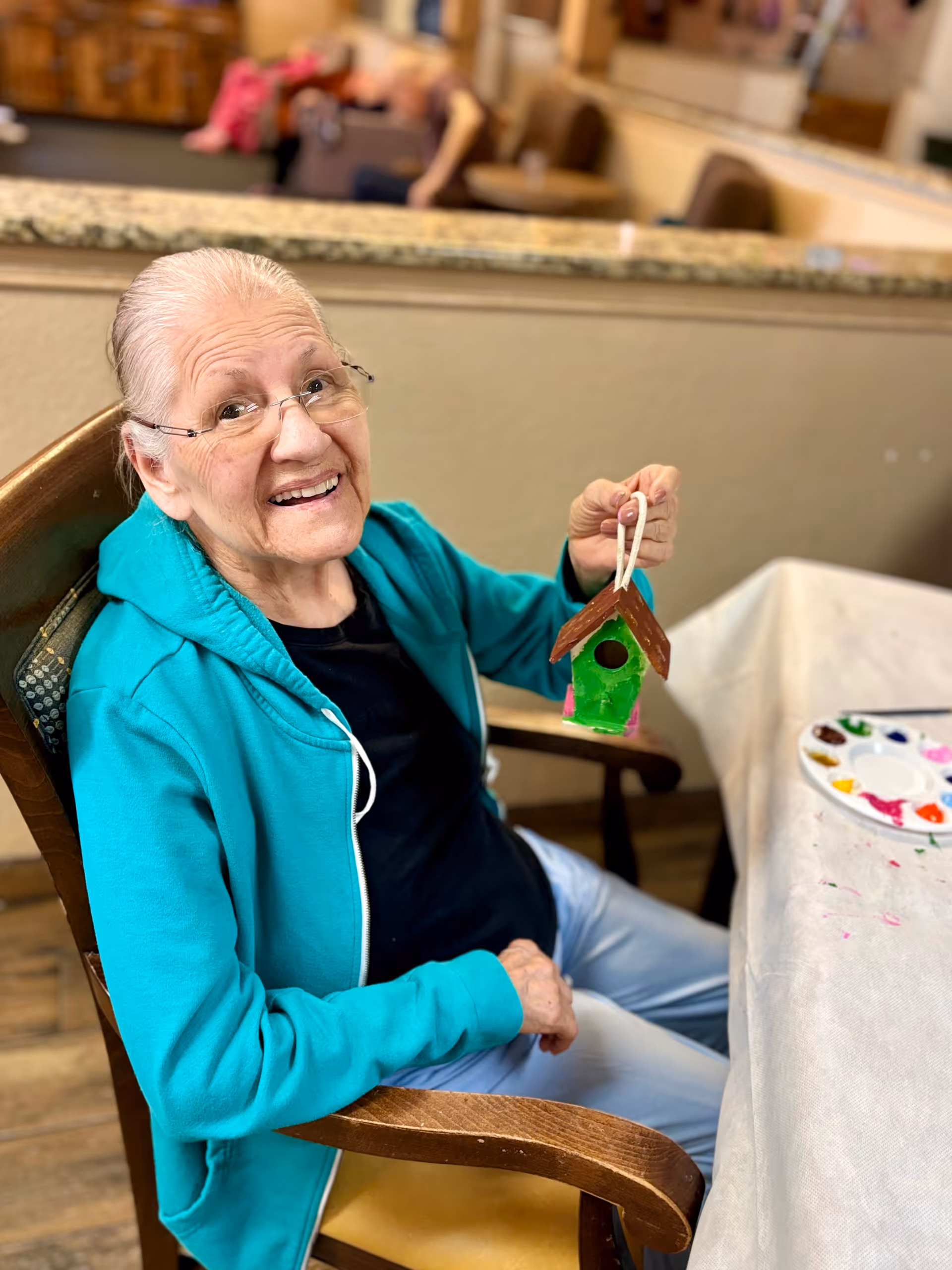 An elderly woman wearing glasses and a teal hoodie is sitting in a wooden chair at a table covered with a white cloth. She is smiling and holding up a small, colorful birdhouse she has painted. A paint palette with various colors is on the table beside her. The background shows a granite countertop and blurred furniture.