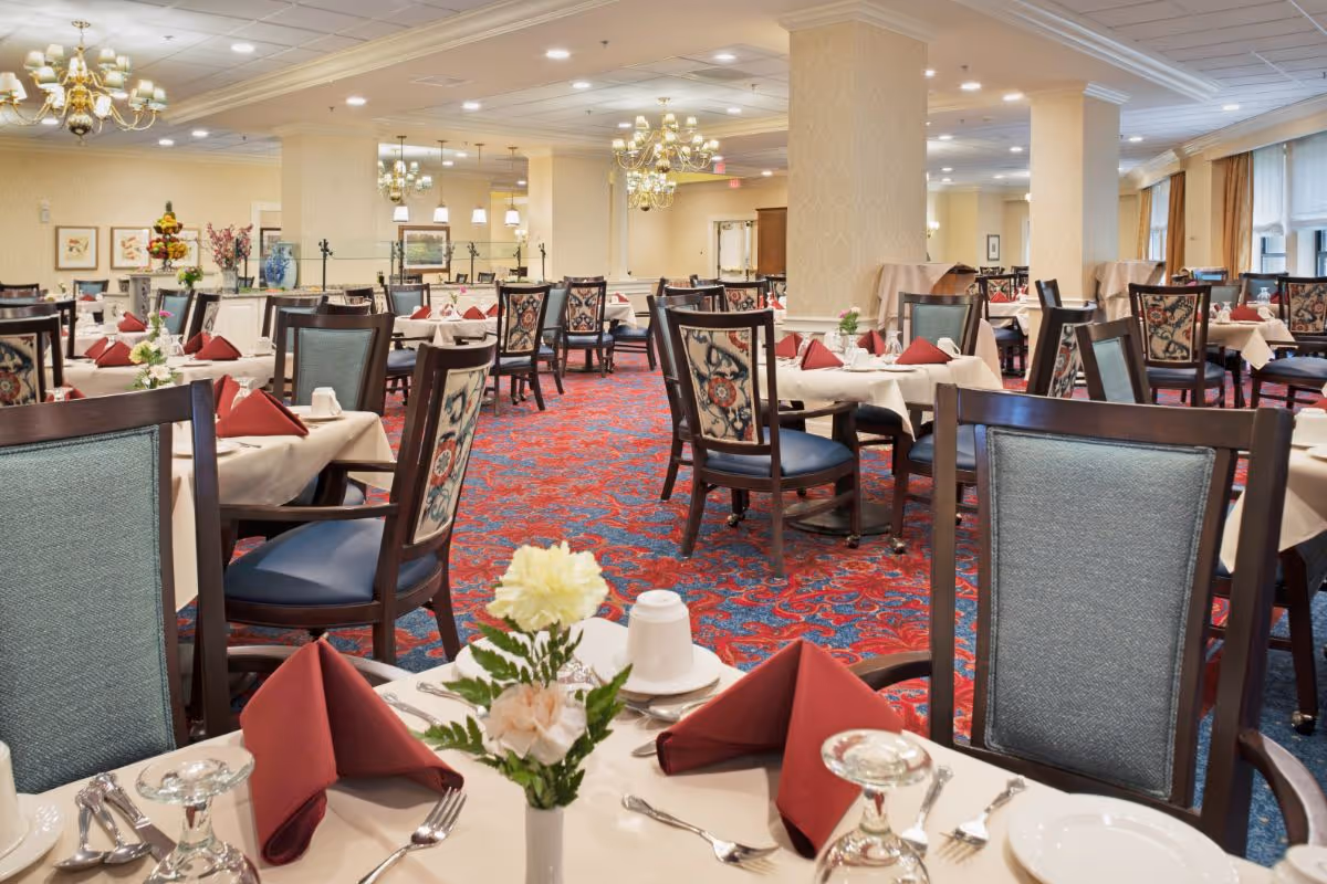 Spacious dining room with neatly set tables, floral centerpieces, and upholstered chairs.
