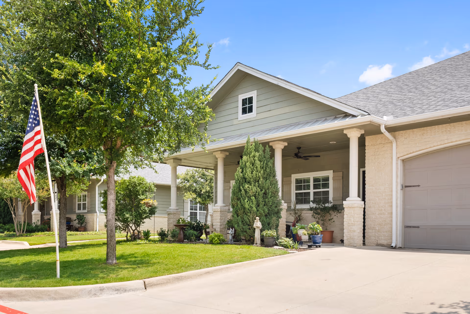 Front exterior of a single-story residential building with a covered porch, columns, potted plants and an American flag on the lawn.