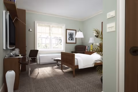 A well-lit bedroom in a senior living facility featuring a single wooden bed with white bedding, a brown armchair with an ottoman, a small desk with a chair, a wall-mounted TV, a window with blinds, and a potted plant near the entrance.