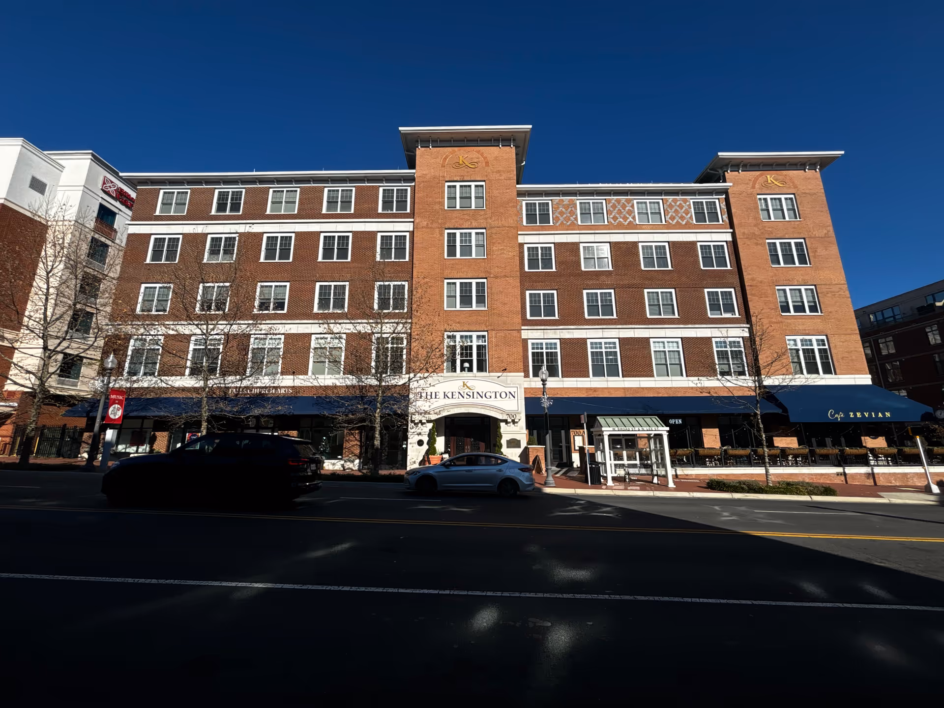 Front view of The Kensington Falls Church, a multi-story brick building with storefront awnings and cars parked on the street.