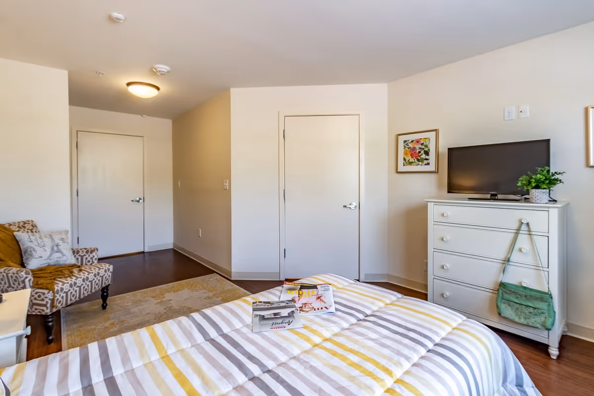 A bright bedroom with a striped bedspread in shades of yellow, gray, and white. There is a white dresser with a small TV and a potted plant on top. A green handbag hangs from one of the dresser knobs. A patterned armchair with a throw blanket and pillow is positioned near the door. The walls are light-colored, and there are two closed doors visible. A framed floral artwork hangs above the dresser.