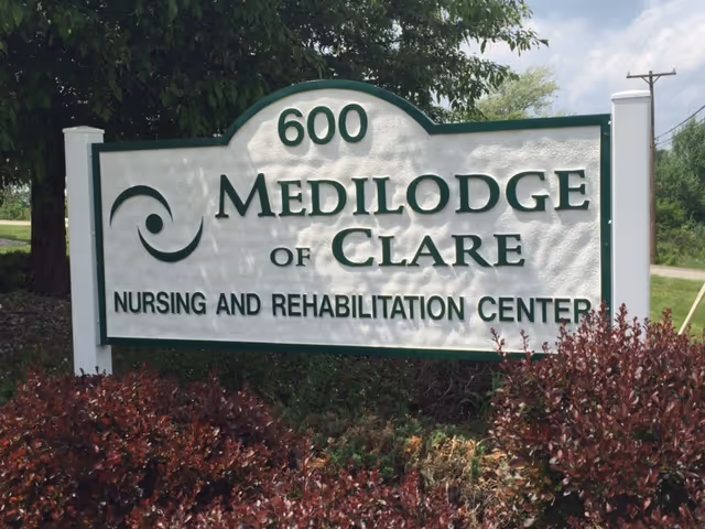 Outdoor sign for Medilodge of Clare Nursing and Rehabilitation Center with the address number 600, surrounded by bushes and trees under a partly cloudy sky.