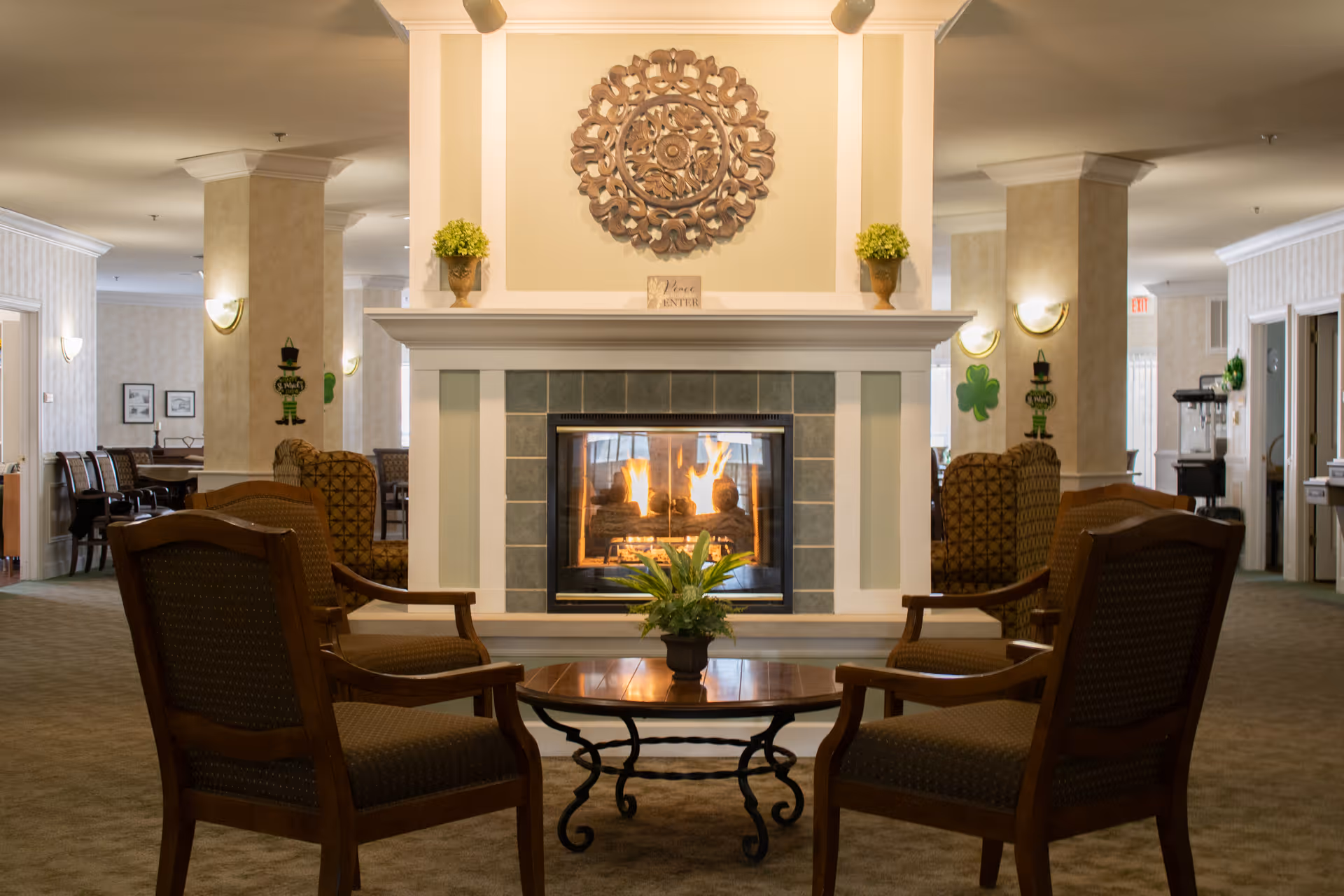 A cozy senior living facility common area with a lit fireplace in the center, surrounded by four wooden armchairs with cushioned seats. A round wooden coffee table with a small potted plant sits in front of the fireplace. The room has beige walls, carpeted floors, and decorative wall sconces. There are columns and additional seating areas visible in the background.