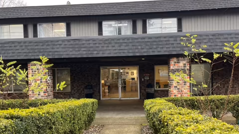 Front entrance of a two-story brick senior living building with a hedged walkway leading to glass double doors.