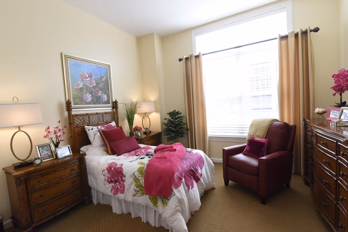 Bright bedroom with a twin bed dressed in pink floral linens, wooden nightstands and dresser, and a burgundy armchair beside a large window.