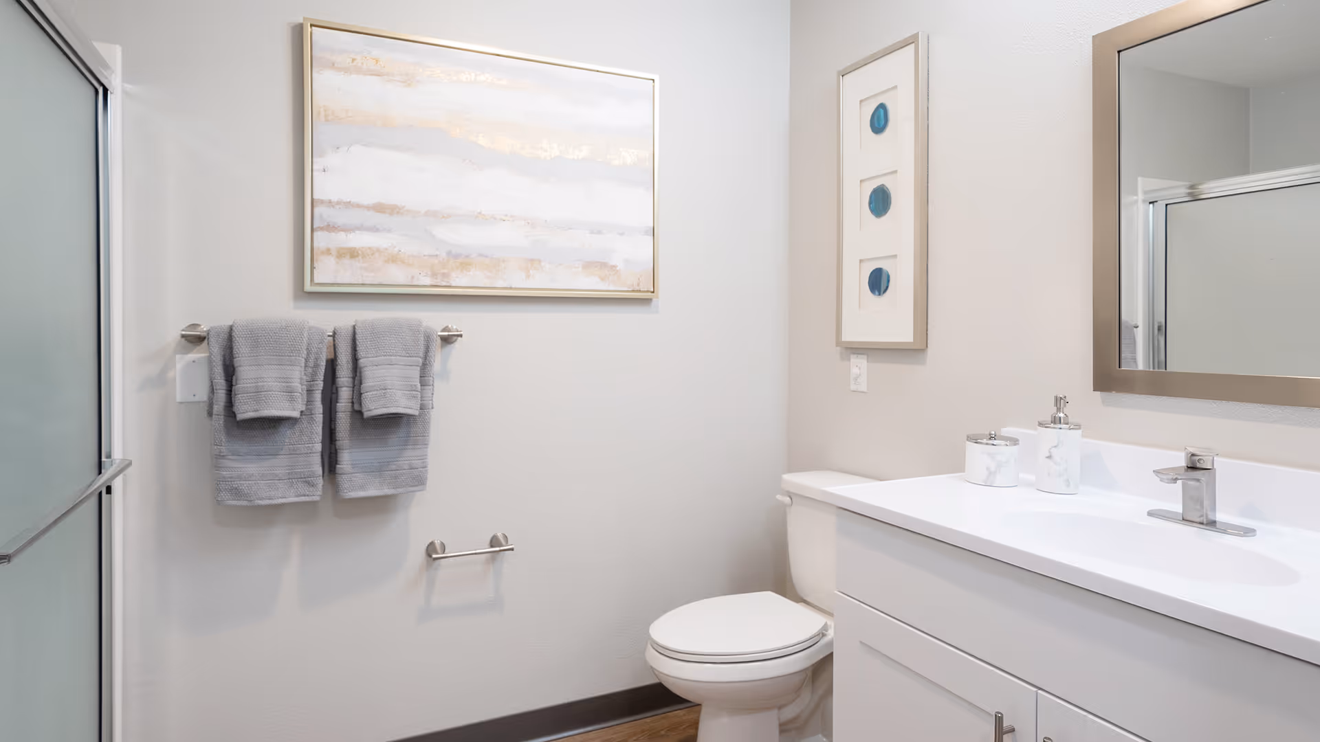 Clean modern bathroom with a white vanity and sink, toilet, towel rack with gray towels, framed artwork, and a shower with frosted glass.