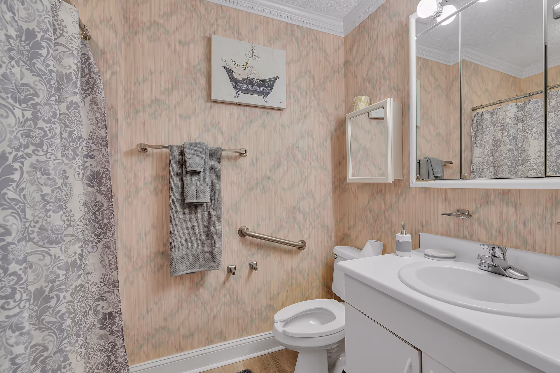 A bathroom with a white sink and countertop, a toilet, a wall-mounted cabinet with a mirror, a towel rack holding gray towels, a patterned shower curtain, and a wall decoration featuring a bathtub illustration.