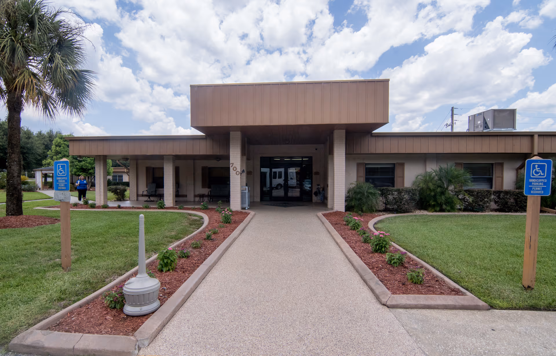 Front entrance of a single-story care facility with a covered drop-off canopy, landscaped walkway, and handicap parking signs.