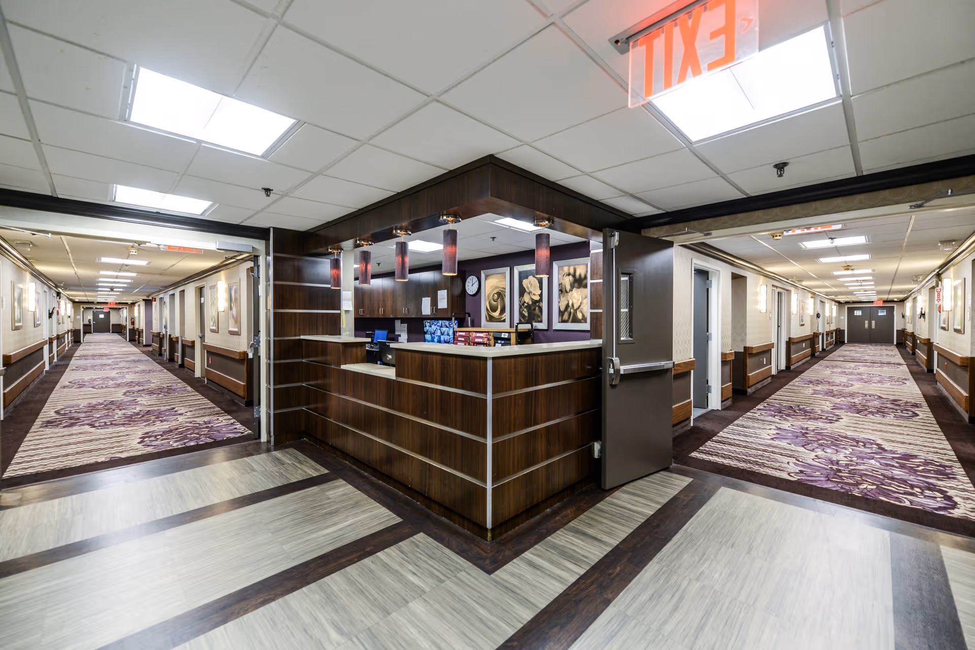 Interior view of a senior living facility hallway with a central nurse's station. The nurse's station has dark wood paneling and purple pendant lights. Two long corridors with patterned purple and beige carpeting extend on either side. The ceiling has recessed lighting and an illuminated exit sign is visible above the nurse's station.