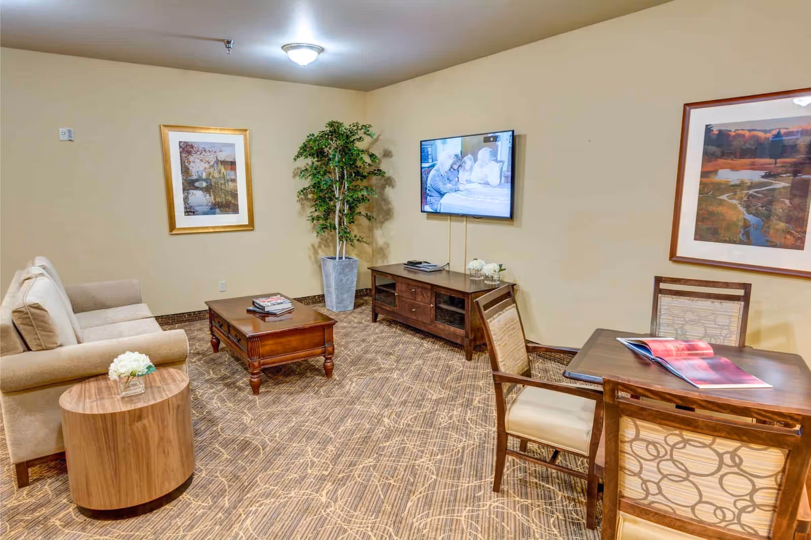 A cozy living room area with a beige sofa, a wooden coffee table with books on it, a round wooden side table with a small flower arrangement, a flat-screen TV mounted on the wall showing an image of three elderly people, a wooden TV stand with decorative items, a potted plant in the corner, and a wooden table with two chairs and an open book. The walls are beige with framed artwork hanging on them, and the floor is carpeted with a patterned design.
